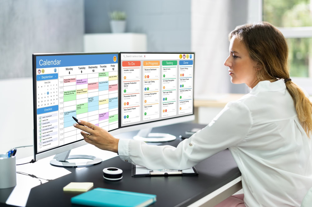 A professional woman in a white blazer is pointing at a large desktop monitor displaying the best content calendar for 2024, in a bright, modern office setting.